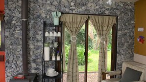 Una habitación con pared de piedra, chimenea, estante con utensilios de cocina, una silla y vistas a través de un ventanal.