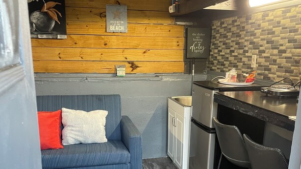 A compact kitchen with a refrigerator, a sink, and a bar area. There's a blue sofa with red and white cushions, and a wooden wall with a framed quote. - Cozy 1-bedroom studio tiny home experience (Orlando)