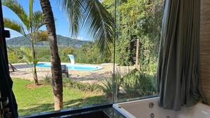 Habitación con amplia ventana que da a una piscina y follaje, bañera y una pequeña mesa con un recipiente de vidrio.