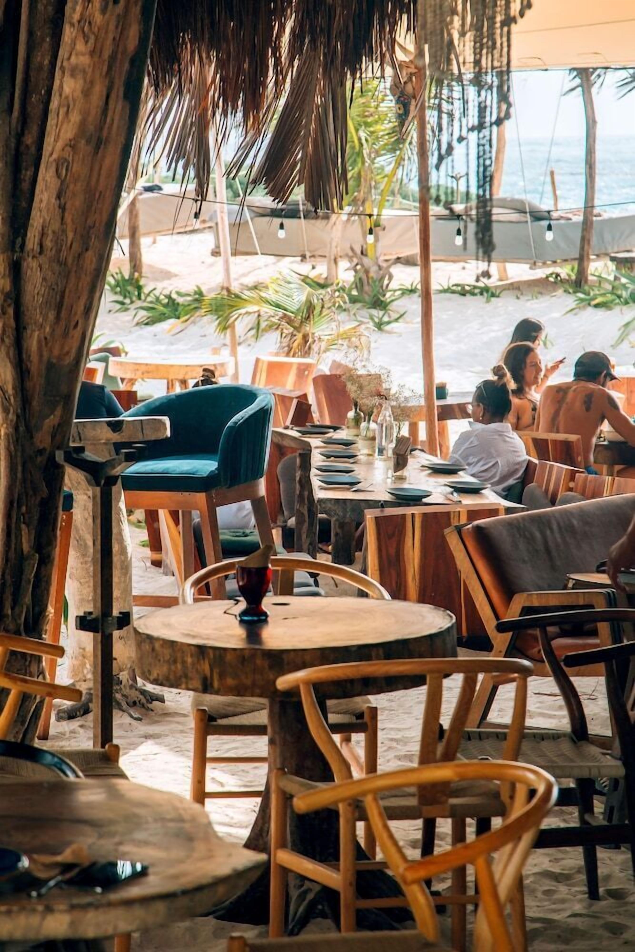 An outdoor dining area with wooden tables and chairs, a thatched roof, and a view of the beach and ocean.