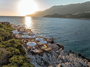En restaurang vid kusten med uteservering och utsikt över havet, en träbrygga och en klippig terräng.