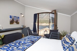 Ein Hotelzimmer mit Bett, einer Sitzecke, einem kleinen Tisch, einem Sessel, einem Balkon mit Meerblick und einem Bild an der Wand.