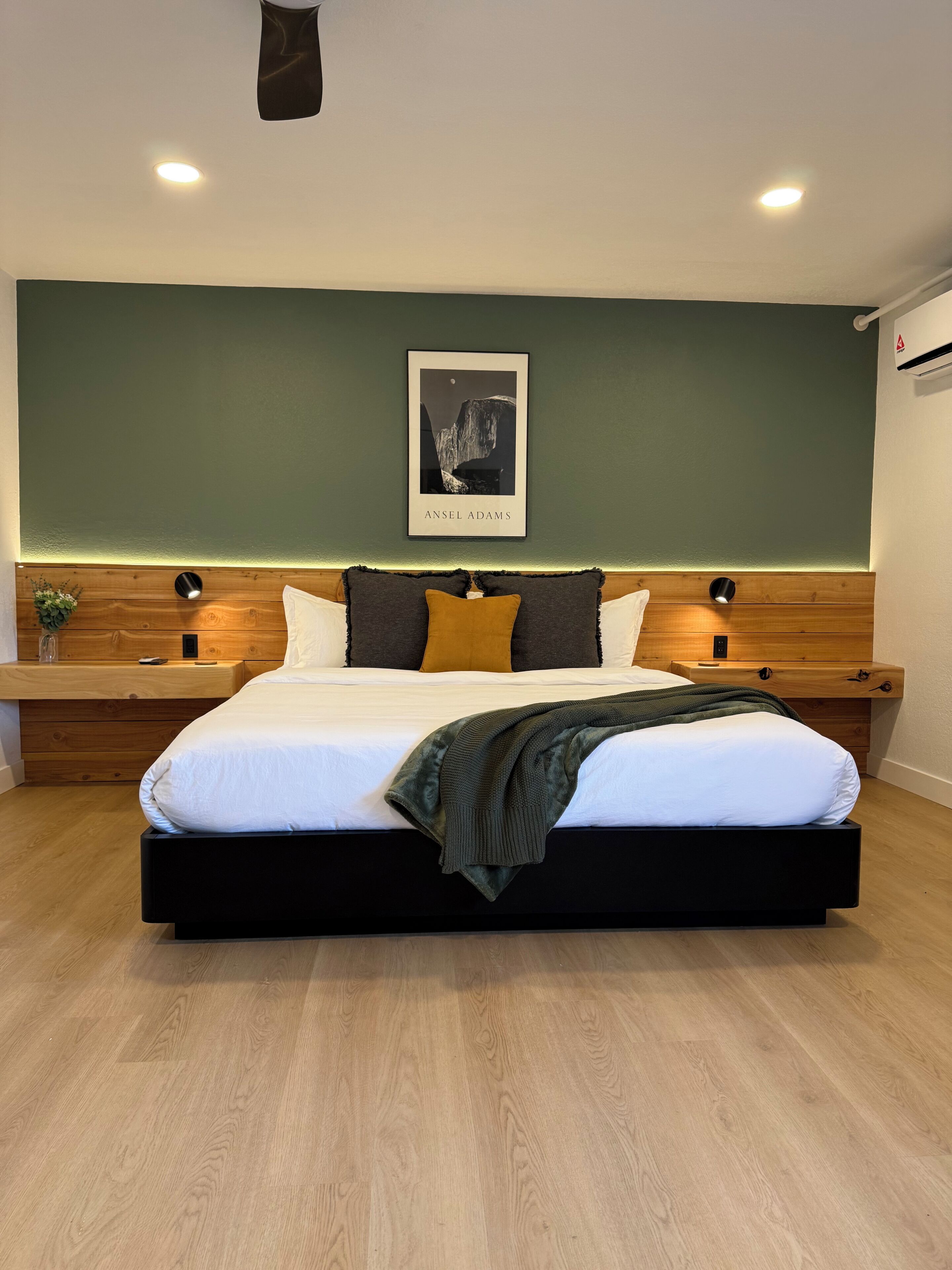 Un dormitorio moderno con una cama grande, cabecera de madera, dos mesitas de noche, una obra de arte enmarcada y una mesita con una planta.