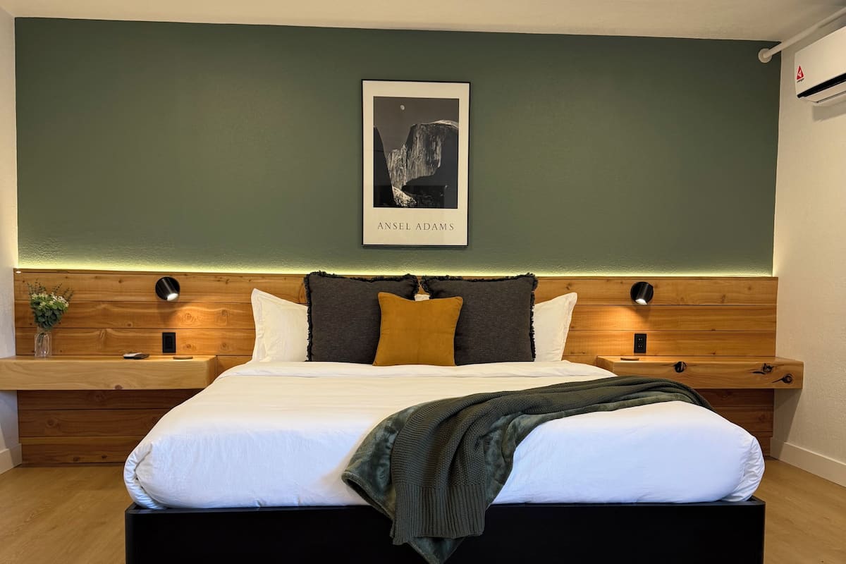 Un dormitorio moderno con una cama grande, cabecera de madera, dos mesitas de noche, una obra de arte enmarcada y una mesita con una planta.