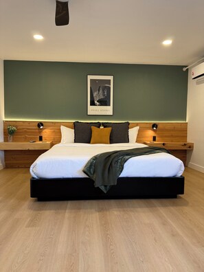 Un dormitorio moderno con una cama grande, cabecera de madera, dos mesitas de noche, una obra de arte enmarcada y una mesita con una planta.