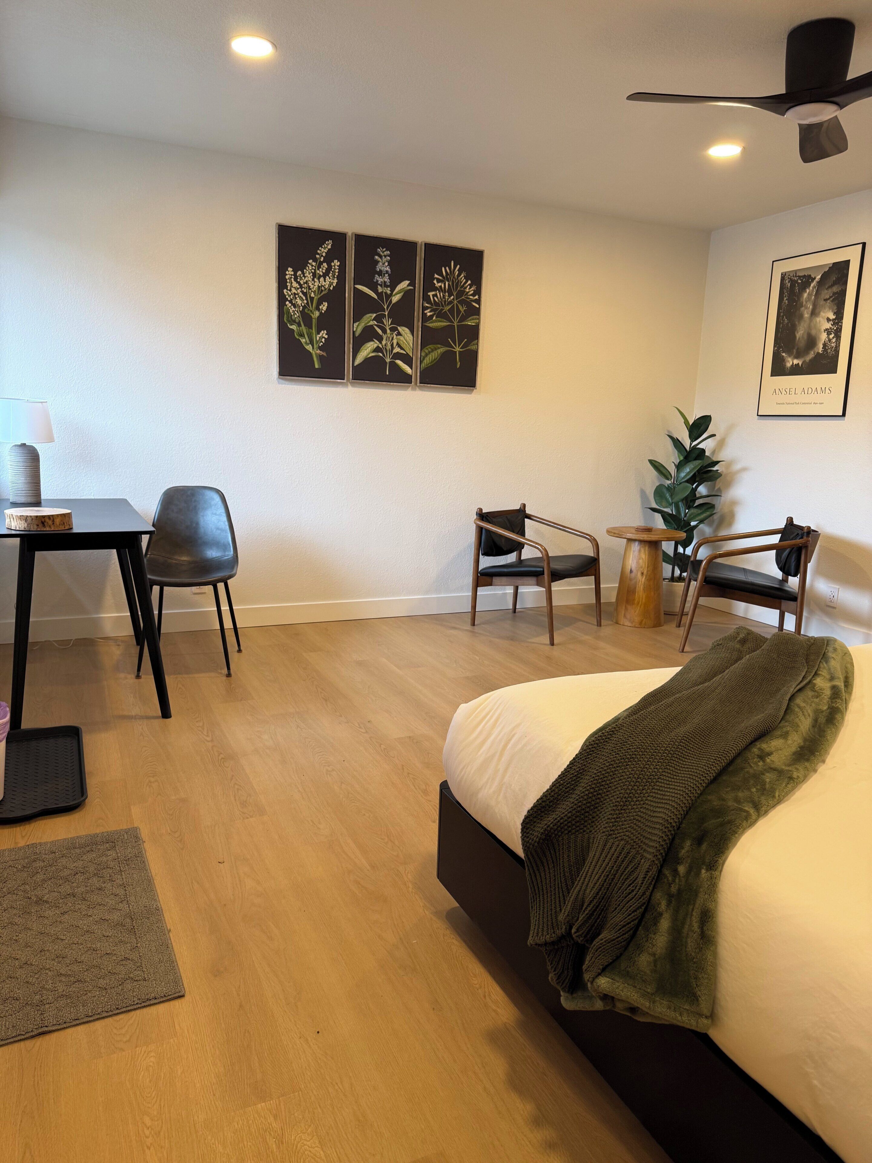 Habitación de hotel con cama, escritorio, dos sillas, una planta y obras de arte en las paredes.