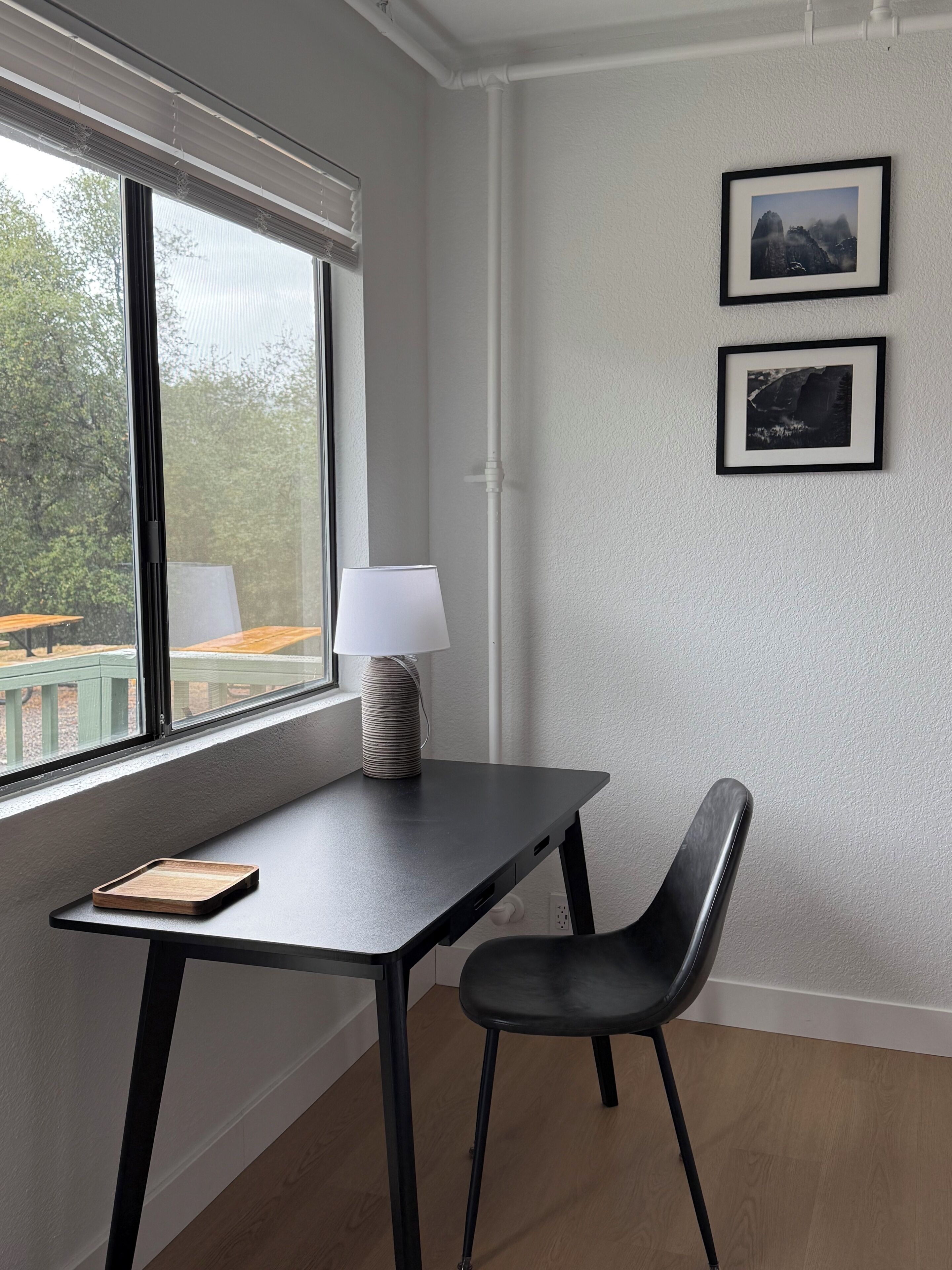 Ein Zimmer mit Schreibtisch, Stuhl, Fenster, Lampe und zwei gerahmten Bildern an der Wand.