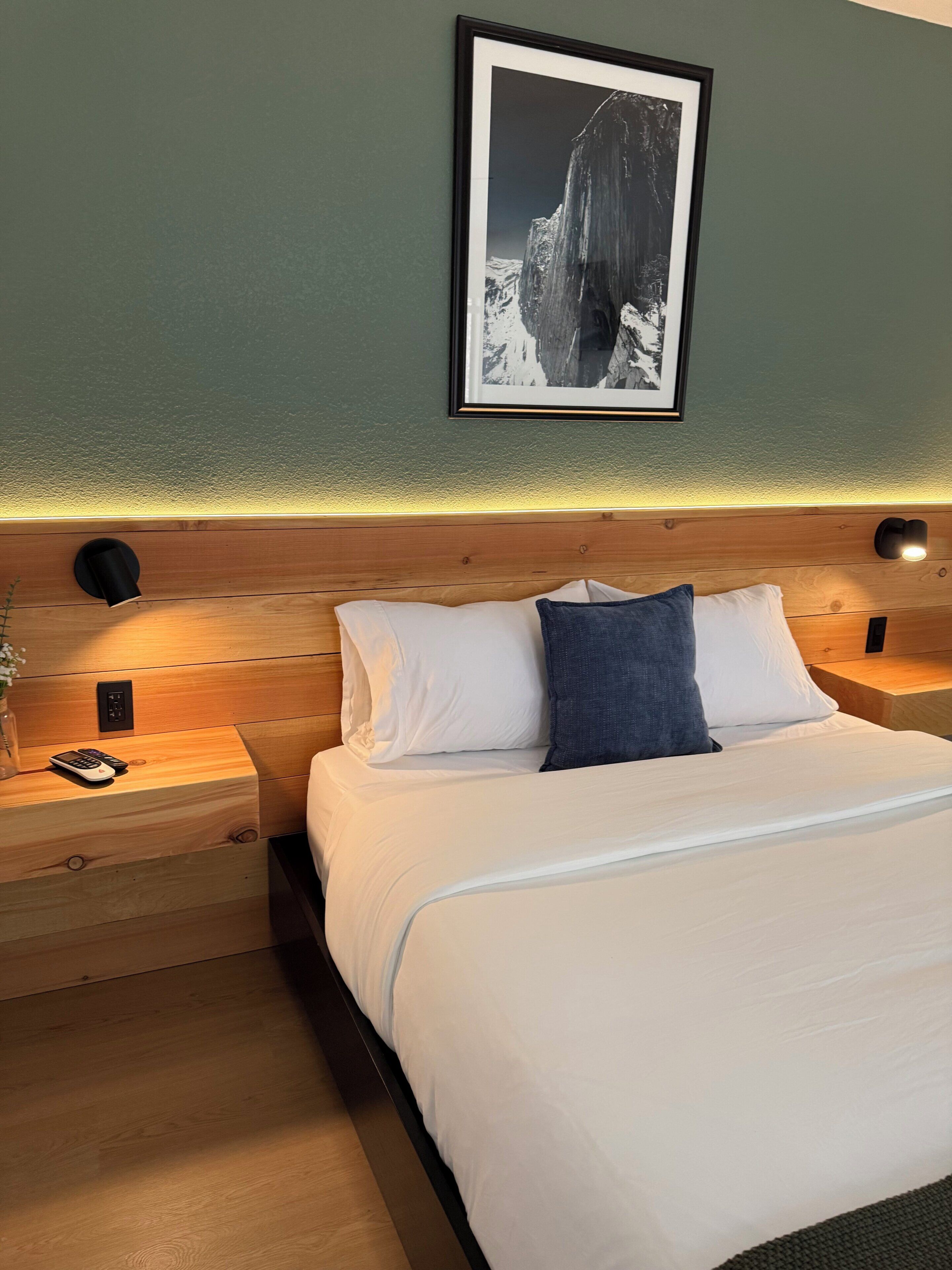 Cama bem arrumada com almofadas brancas e azuis, cabeceira de madeira, mesas de cabeceira com abajures e um quadro na parede.