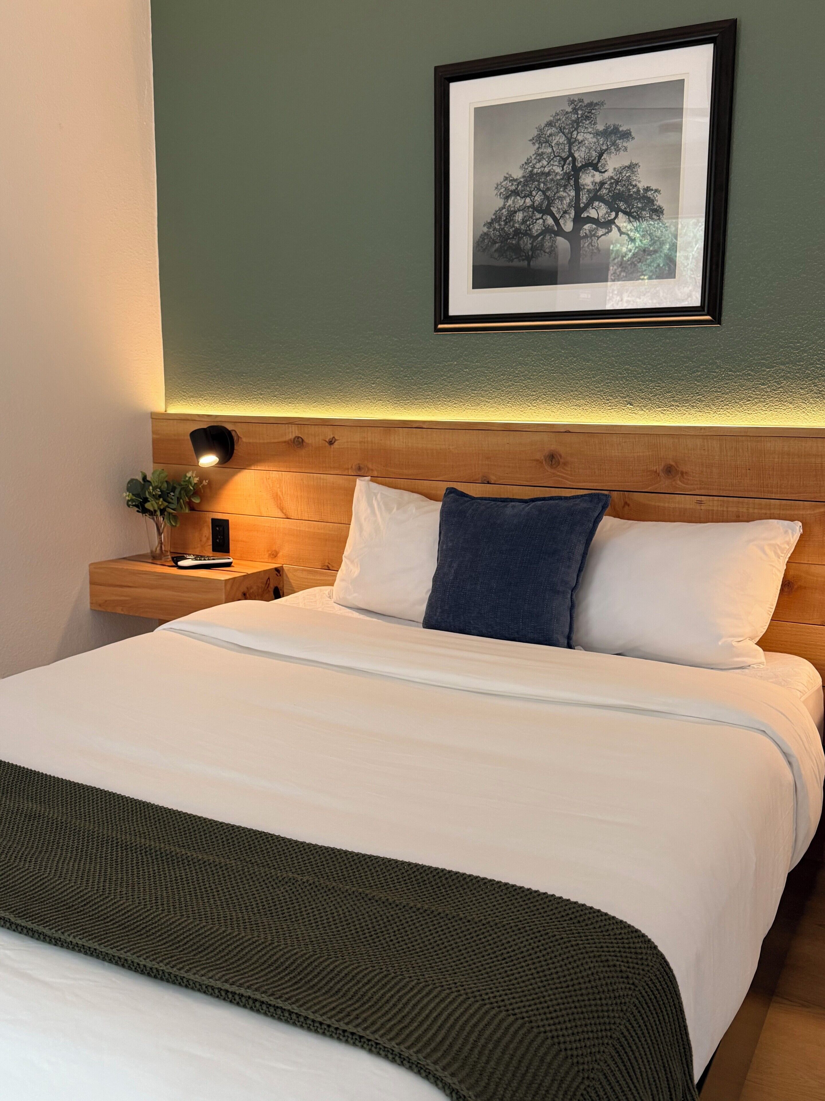 A neatly made bed with white and blue pillows, a wooden headboard, a nightstand with a lamp, and a framed picture on the wall.