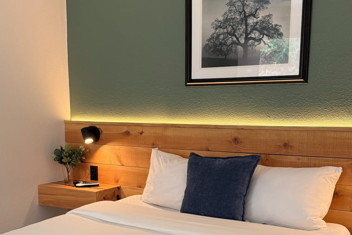 A neatly made bed with white and blue pillows, a wooden headboard, a nightstand with a lamp, and a framed picture on the wall.