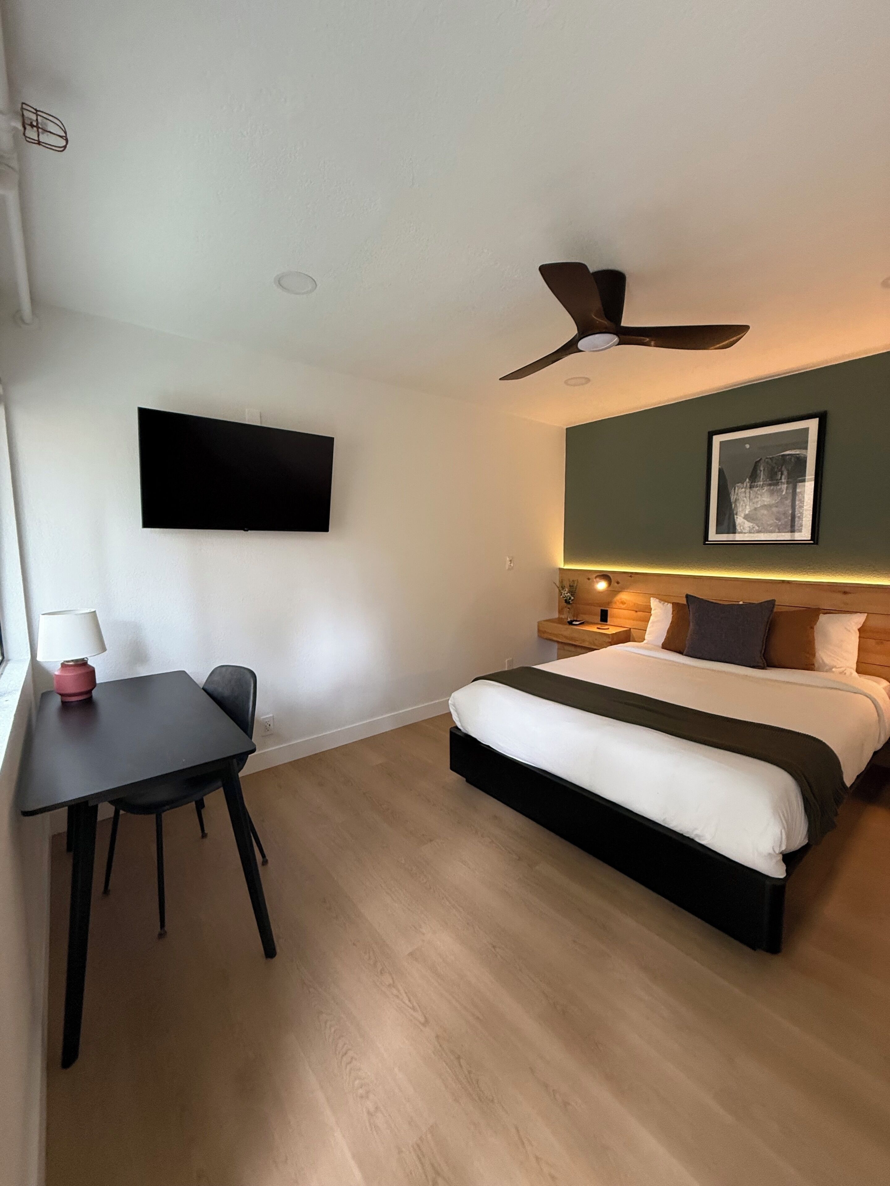 Ein Schlafzimmer mit einem Bett, einem Schreibtisch, einem Stuhl, einem Deckenventilator, einem Fernseher und einem gerahmten Bild an der Wand.