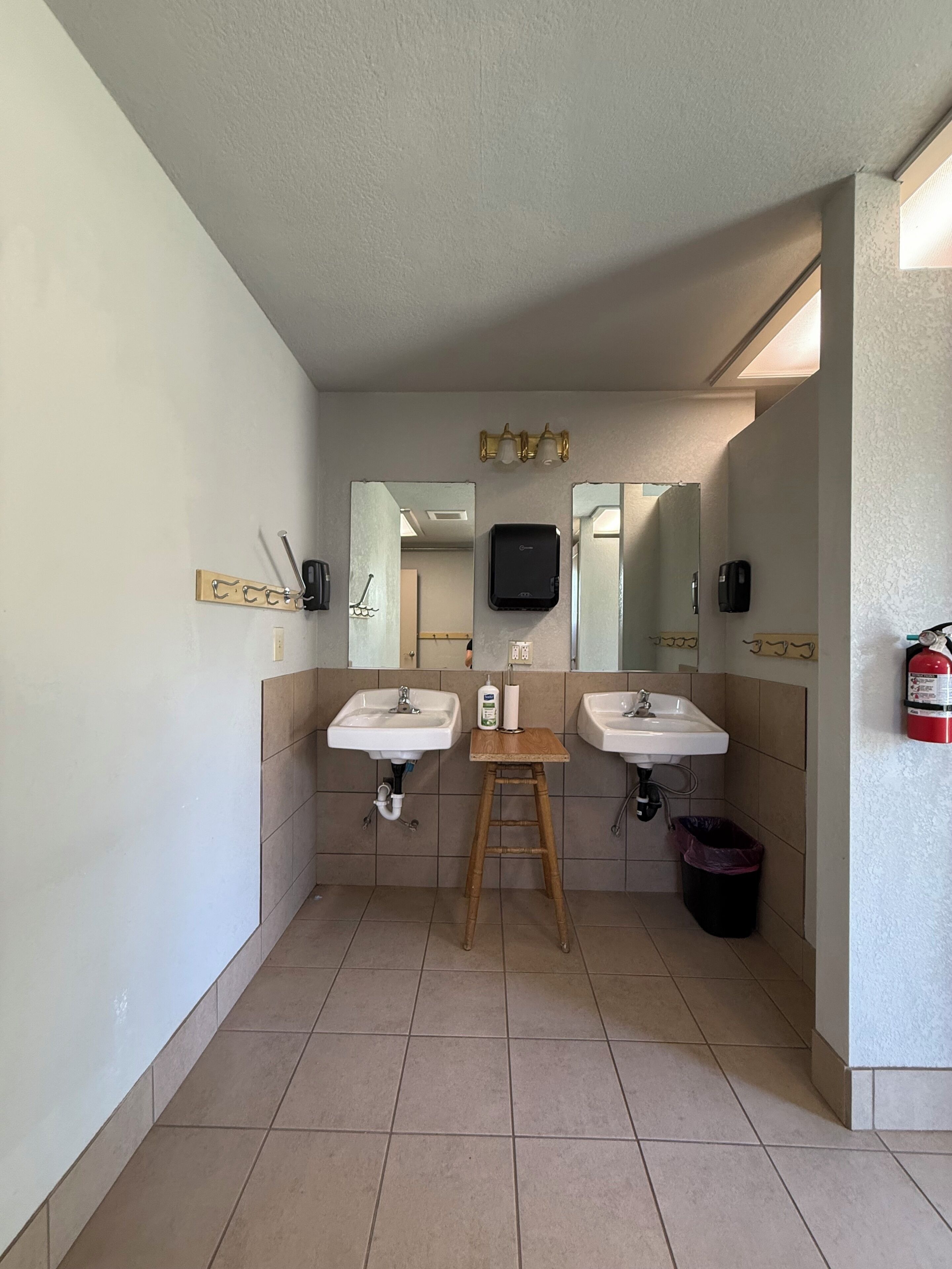 Un baño con dos lavamanos, un taburete de madera, un espejo y un extintor.