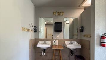 Un baño con dos lavamanos, un taburete de madera, un espejo y un extintor.
