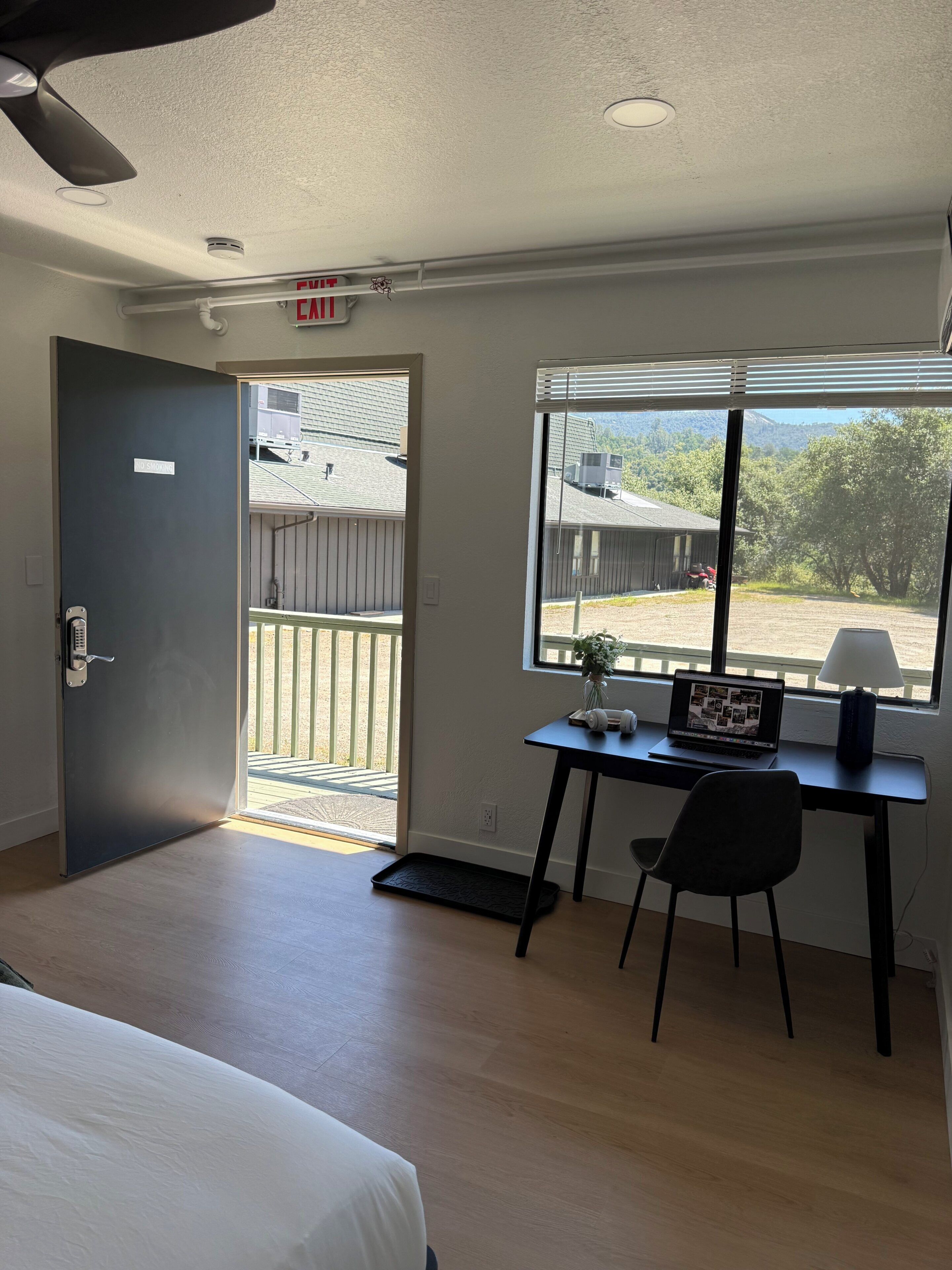 A hotel room with a bed, a desk with a laptop, a chair, a window with a view of buildings and trees, and a door with an exit sign.