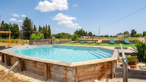 Una piscina rectangular de madera con un deck de madera alrededor, una pérgola y un área verde con sillas de descanso.