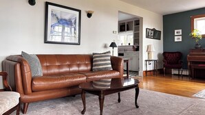 Uma sala de estar com um sofá de couro marrom, uma mesa de centro de madeira, uma poltrona vermelha, um piano e uma área de cozinha ao fundo.