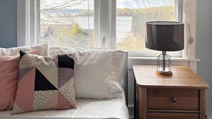 Quarto com cama, almofadas, mesa de cabeceira, abajur e duas janelas com vista para o exterior.