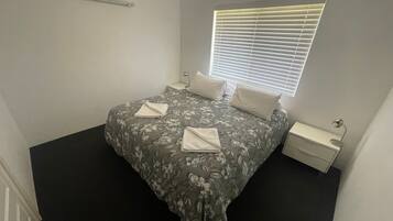 A neatly made bed with a floral bedspread, two pillows, and two folded towels. There is a bedside table with a lamp, a window with blinds, and a small nightstand with a lamp.