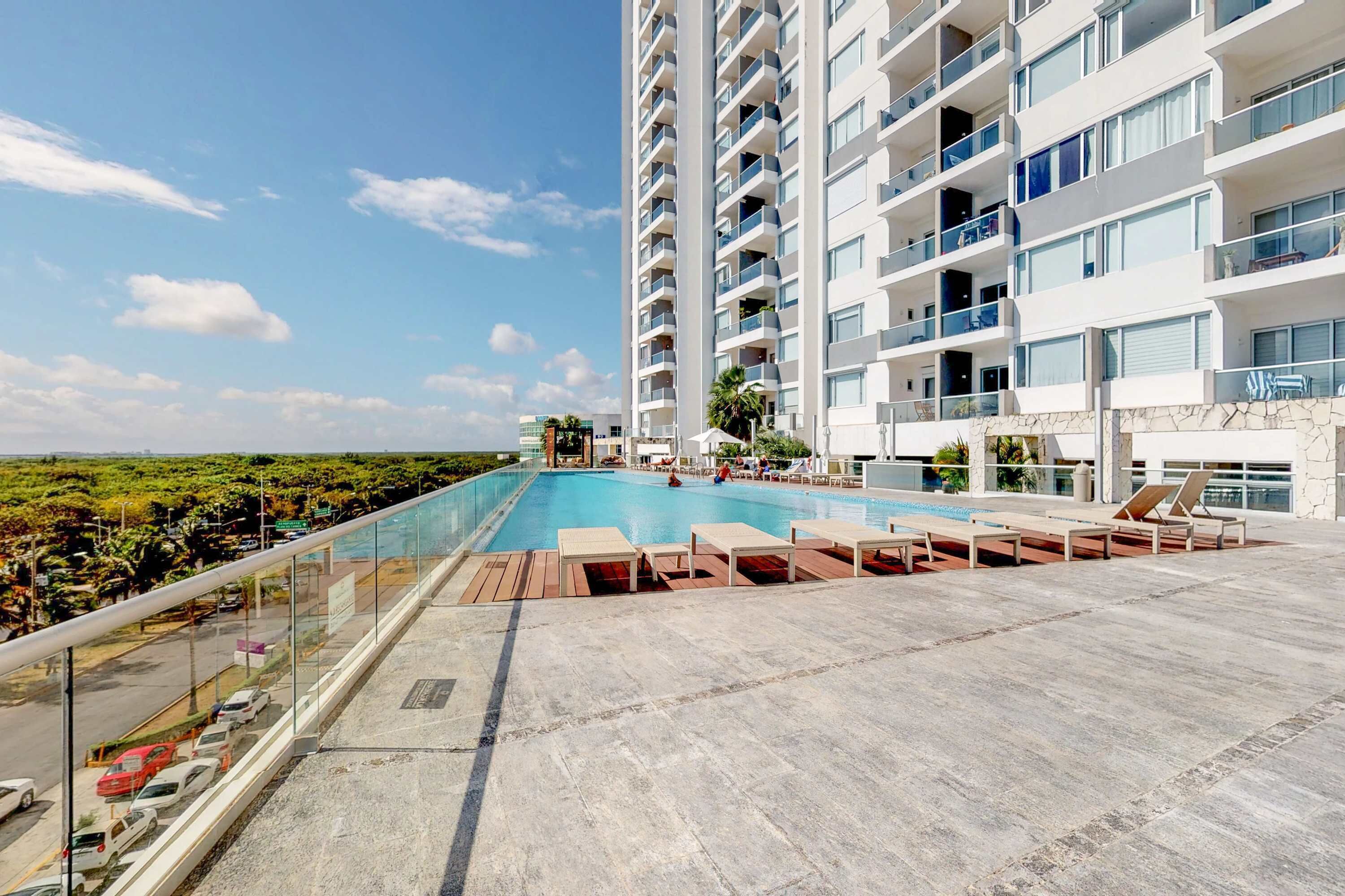 A rooftop pool with lounge chairs, a glass balustrade, and a high-rise building with balconies.