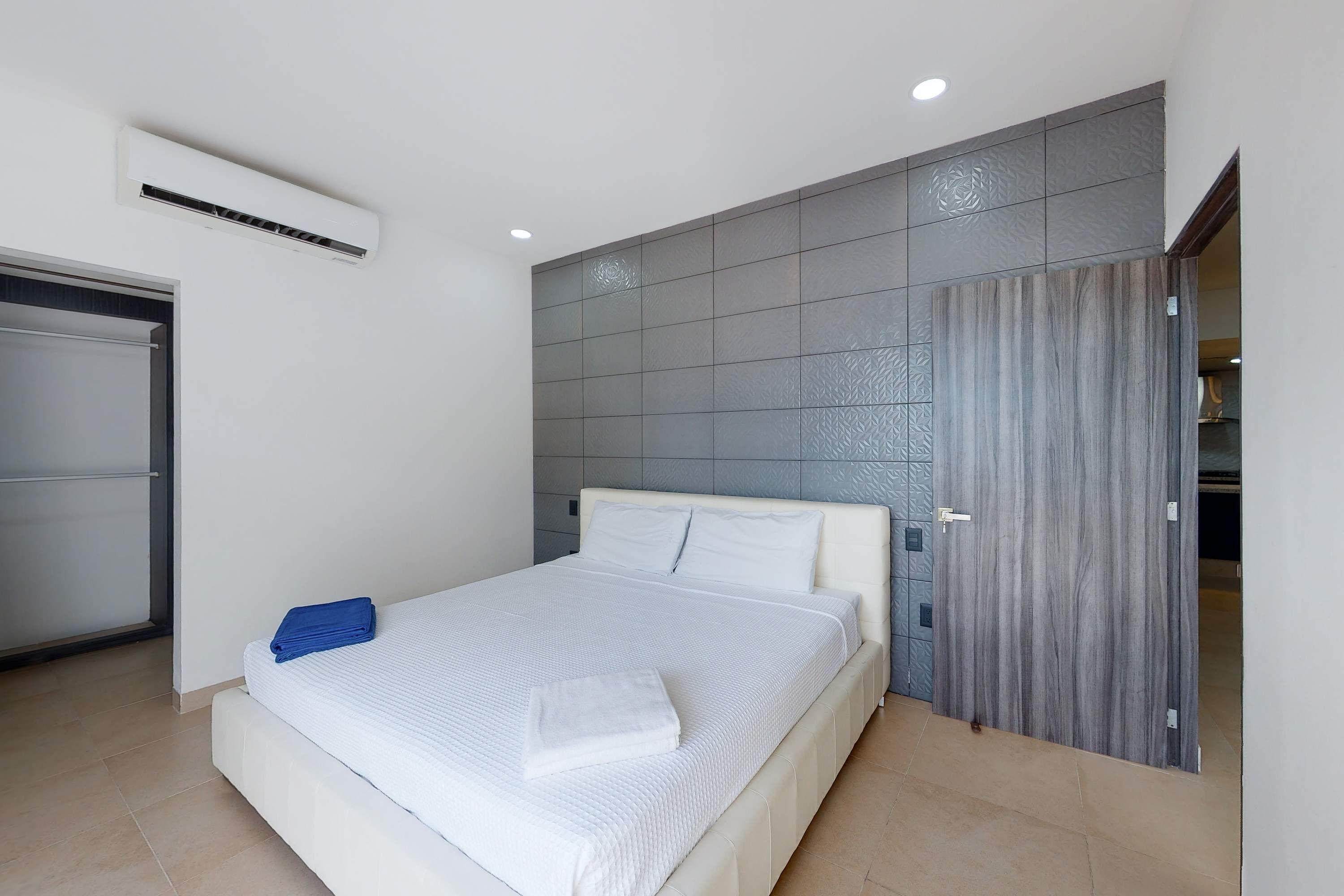 A bedroom with a bed, a white air conditioner, a grey textured wall, and a door.
