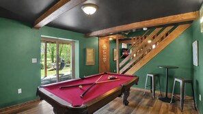 Mesa de billar con bolas, una escalera y una zona de bar en una habitación con paredes verdes.