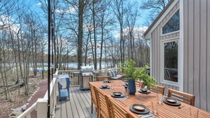 Eine Terrasse mit Esstisch, Grill und Blick auf einen von Bäumen umgebenen See.