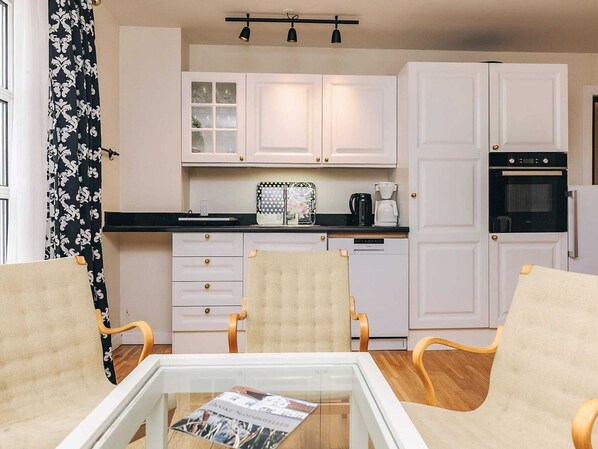 A compact kitchen with white cabinets, a coffee maker, and a refrigerator. There are two chairs and a table with a magazine.