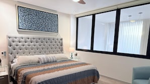 Un dormitorio con cabecera capitonada, un ventanal con cortinas, una mesita de noche con lámpara y una cama con colcha a rayas.