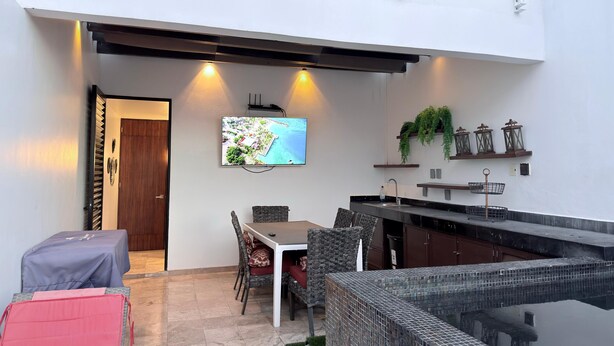 Un área de comedor exterior moderna con televisor, cocina y piscina.