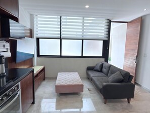 Ein modernes Wohnzimmer mit einer Couch, Hocker, Küchenbereich und großen Fenstern.