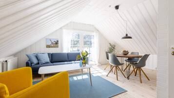 A cozy loft-style room with a high ceiling, a blue sofa, a yellow armchair, a dining table with chairs, and a large window providing natural light.
