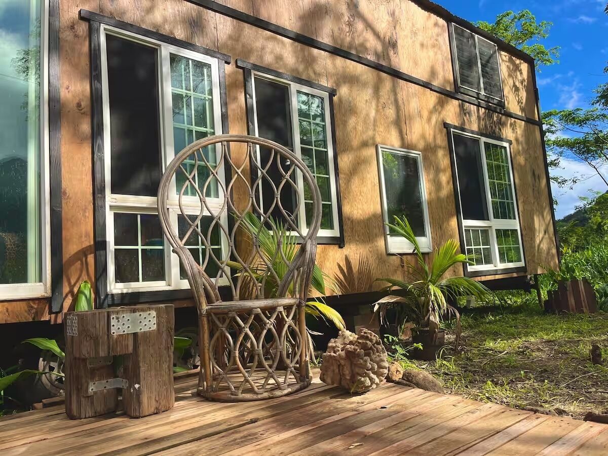 A wooden deck with a wicker chair and a small table, a house with large windows, and a garden with plants and a tree.
