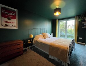A bedroom with a bed, bedside table, lamp, dresser, and a framed picture of Campbell's Tomato Soup. - Cosy and Stylish 1BD Flat in Central Margate (Kent)