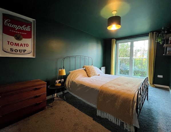 Ein Schlafzimmer mit einem Bett, Nachttisch, Lampe, Kommode und einem gerahmten Bild von Campbell's Tomato Soup.