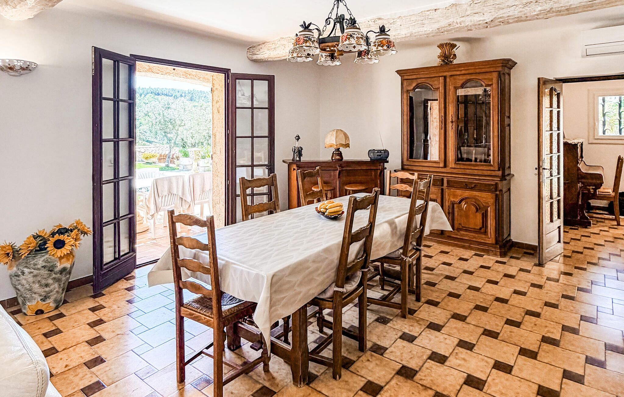 Una sala da pranzo con pavimento a scacchi, sedie di legno, un tavolo con tovaglia bianca, un lampadario e una credenza con vetrate.