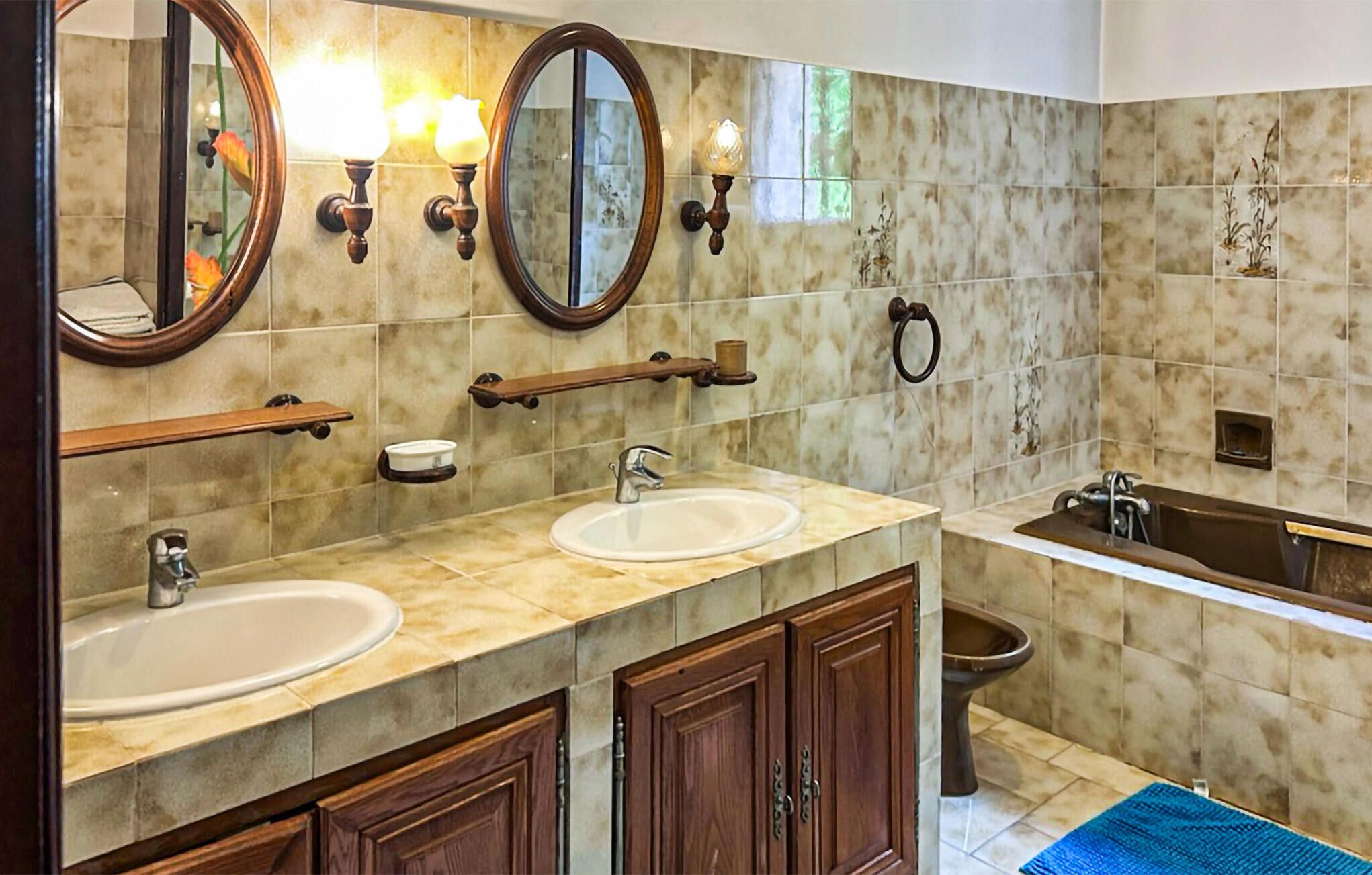 A bathroom with two sinks, a bathtub, and a toilet. There are wooden cabinets and a mirror with wall sconces.