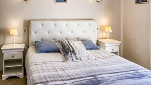 Un dormitorio con cabecera capitonada, dos mesitas de noche con lámparas, una cama con manta a rayas y una mesita de noche con cajón.