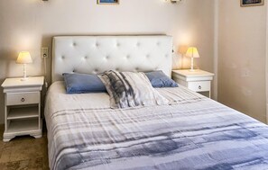 Ein Schlafzimmer mit einem geblatteten Kopfteil, zwei Nachttischen mit Lampen, einem Bett mit gestreifter Bettdecke und einem Nachtschränkchen mit Schublade.