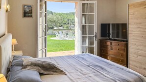 Un dormitorio con la puerta abierta que da a un jardín, una cama con manta, un aparador y un televisor.