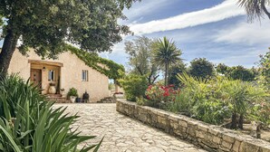 Un sendero de piedra conduce a un edificio rústico con puerta y ventanas, rodeado de exuberante vegetación y plantas con flores.