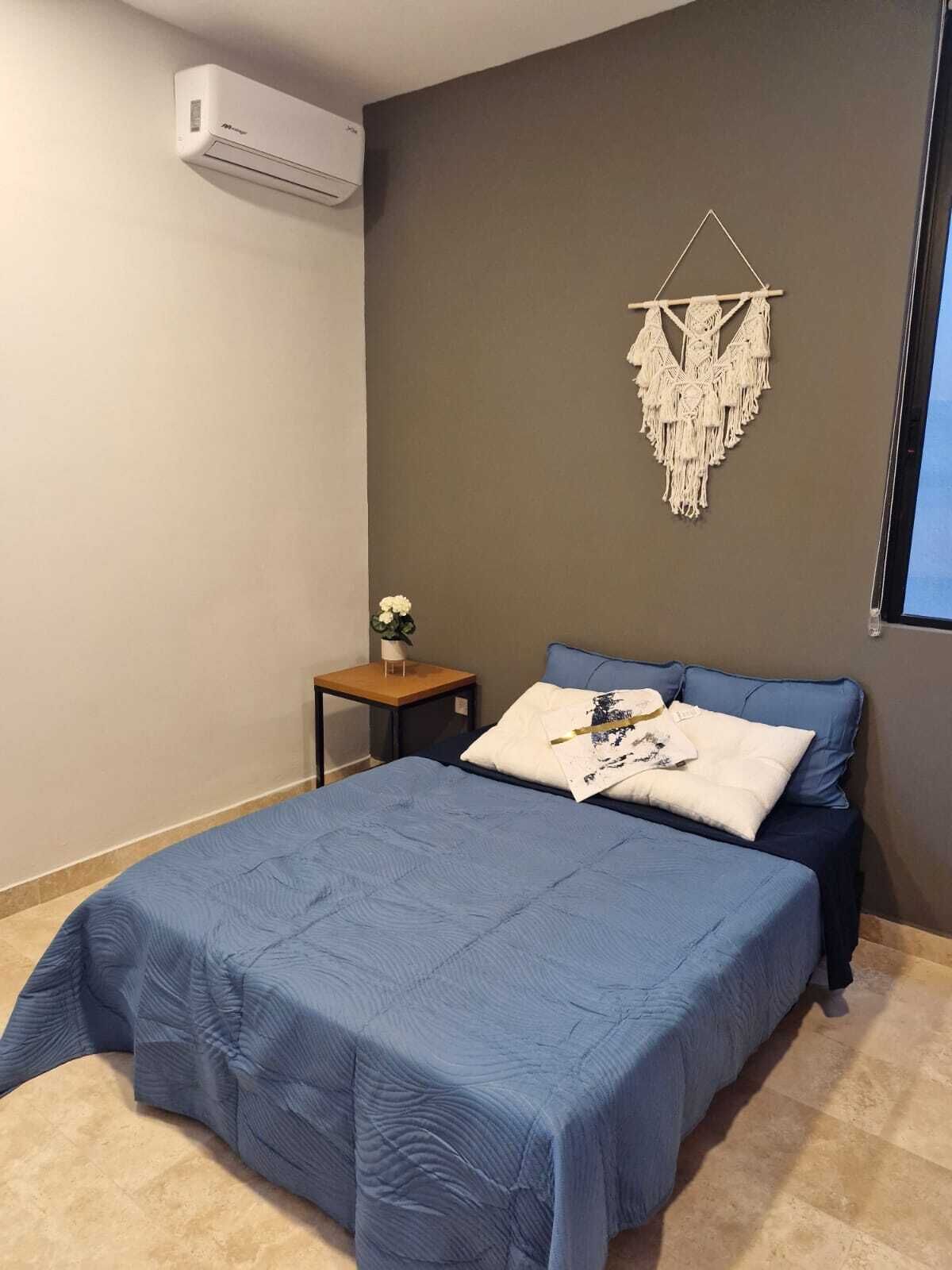 A bedroom with a bed, a small bedside table, a wall-mounted air conditioner, and a macrame wall hanging.