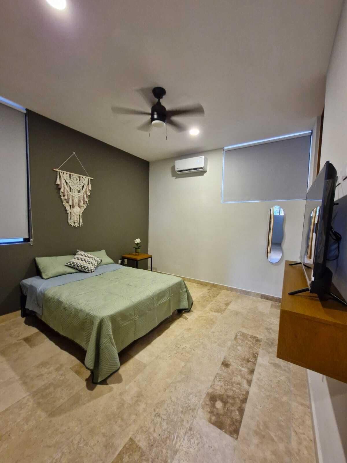 Een slaapkamer met een bed, een plafondventilator, een airconditioning, een klein tafeltje met een vaas en een tv op een standaard.