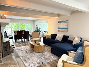 Ein Wohnzimmer mit einem dunkelblauen Sofa, einem Holztisch, einem Essbereich mit Tisch und Stühlen sowie einem großen Fenster mit Blick auf Grünflächen.