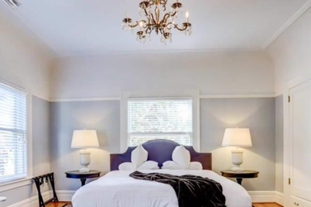Une chambre à coucher comprenant un lit, deux tables de chevet, un banc, un lustre et une fenêtre avec des stores.
