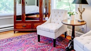 Un dormitorio con un armario de madera, un sillón blanco, una mesa redonda con lámpara y una alfombra estampada.