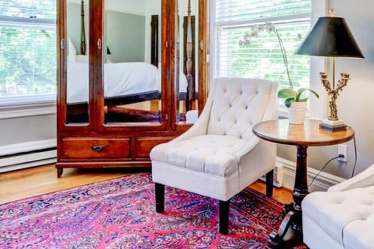 Une chambre à coucher avec une armoire en bois, un fauteuil blanc, une table ronde avec une lampe et un tapis à motifs.