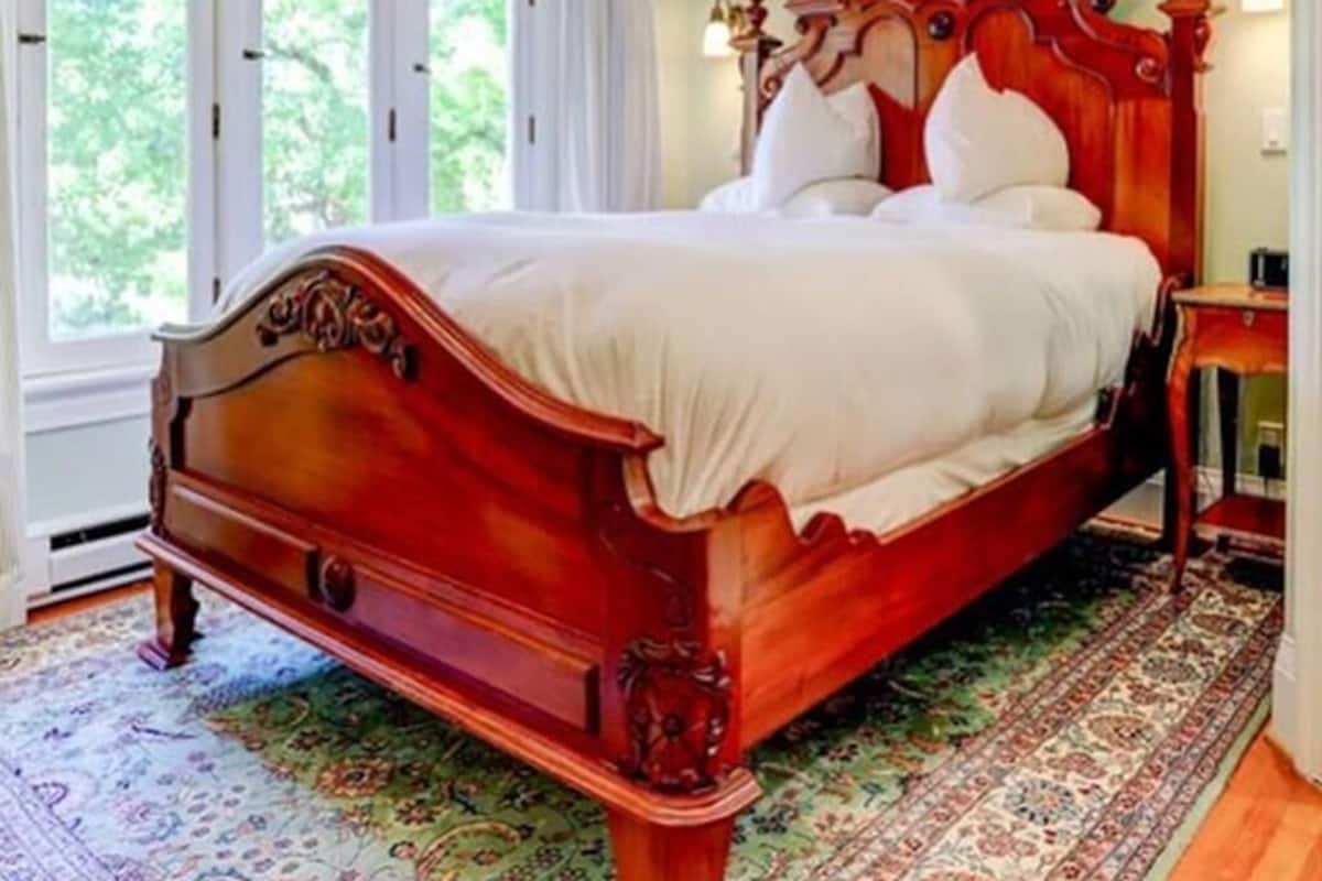 Un lit traditionnel en bois, doté d’une haute tête de lit, d’un tapis à motifs et d’une fenêtre avec des rideaux.