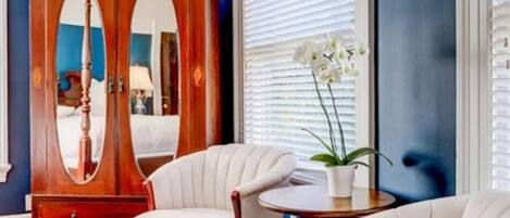 Una habitación con un armario de madera grande, dos sillones blancos tapizados, una mesita redonda y una alfombra estampada.