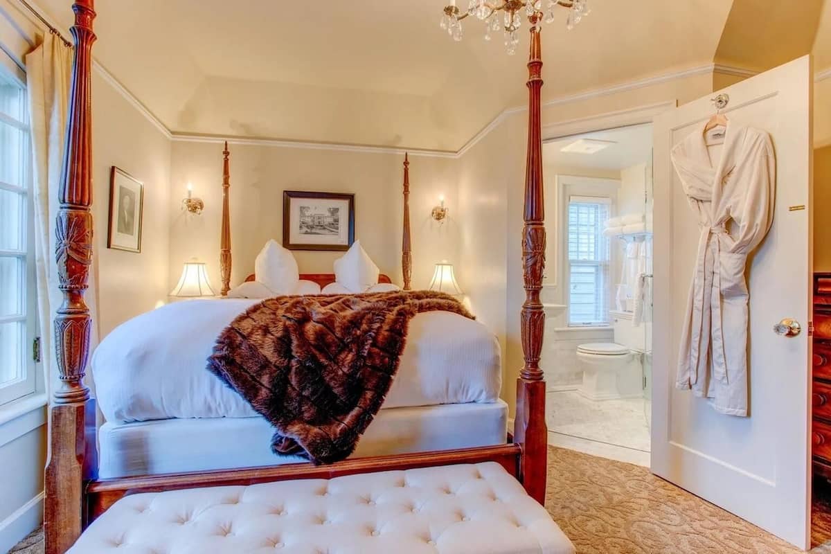 Un lit à baldaquin avec un jeté en fourrure, un lustre et une robe suspendue près de la porte.