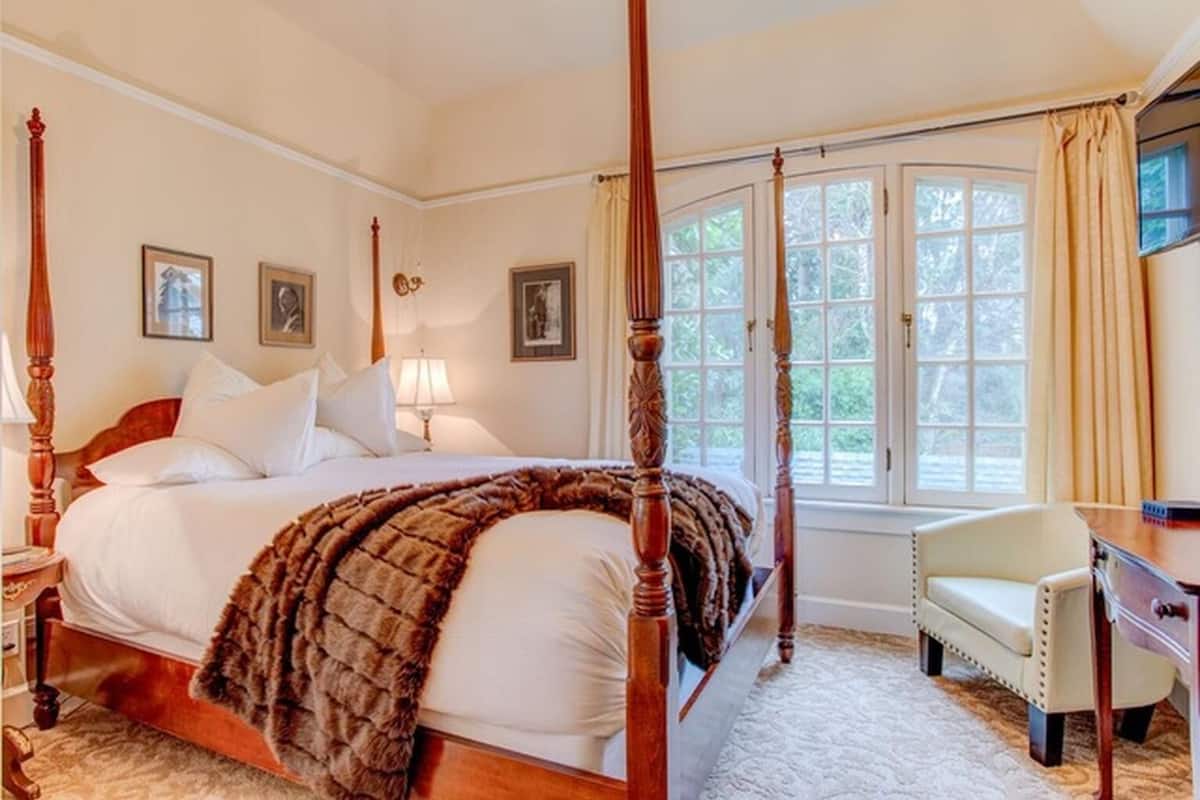 Un lit à baldaquin avec un plaid en fourrure, une table de chevet, une lampe et une chaise dans une pièce dotée de grandes fenêtres et de tableaux encadrés accrochés au mur.